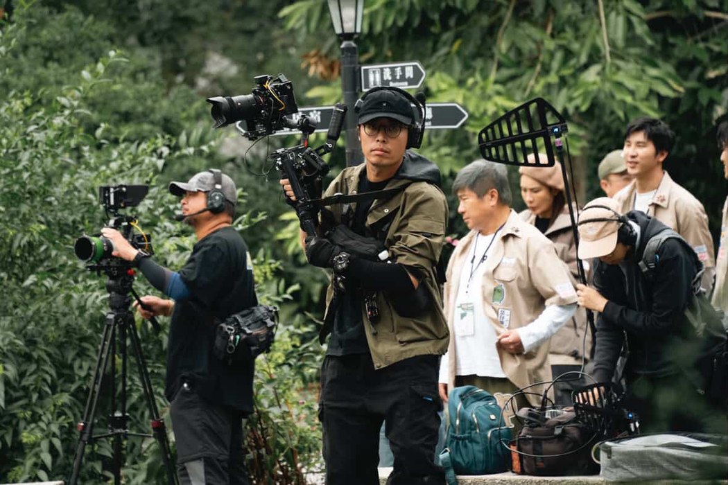 戶外綠地中，攝影與收音工作者帶著器材，和其他參與者一起移動。